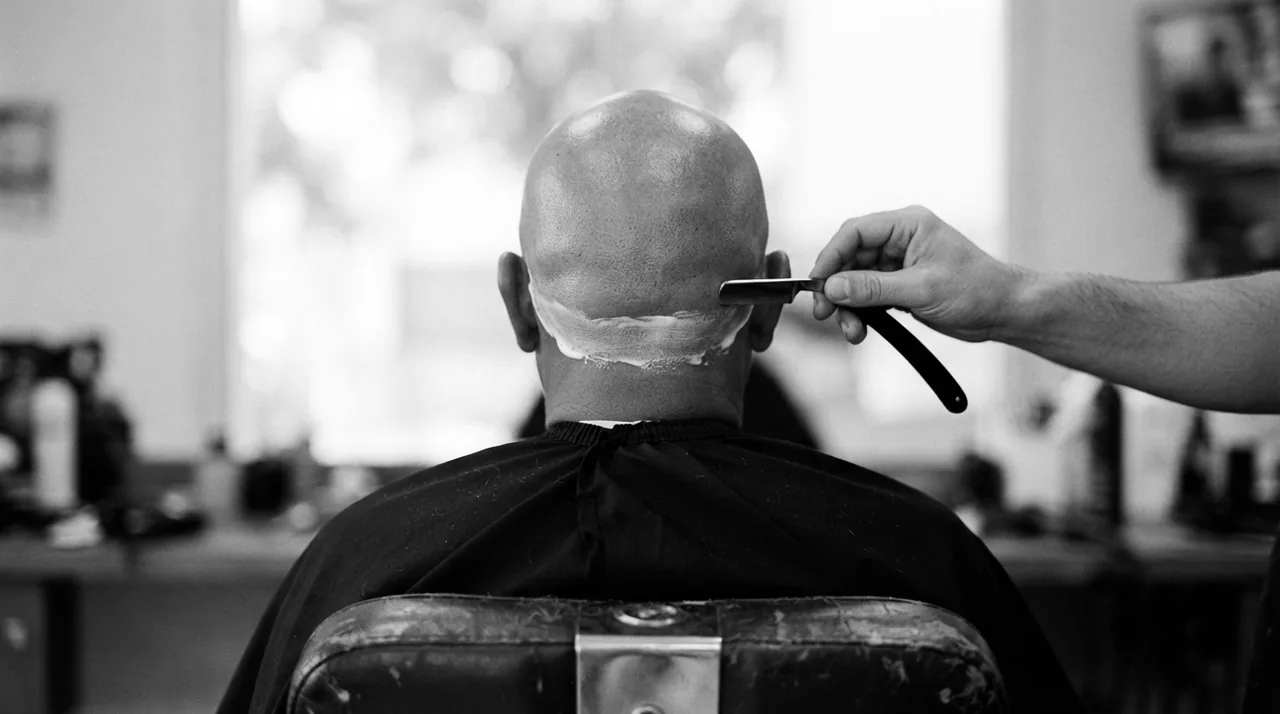 Homme au crâne rasé vu de dos dans un fauteuil de barbier ; la main du barbier tient un rasoir manuel près d'une dernière bande de mousse sur la nuque