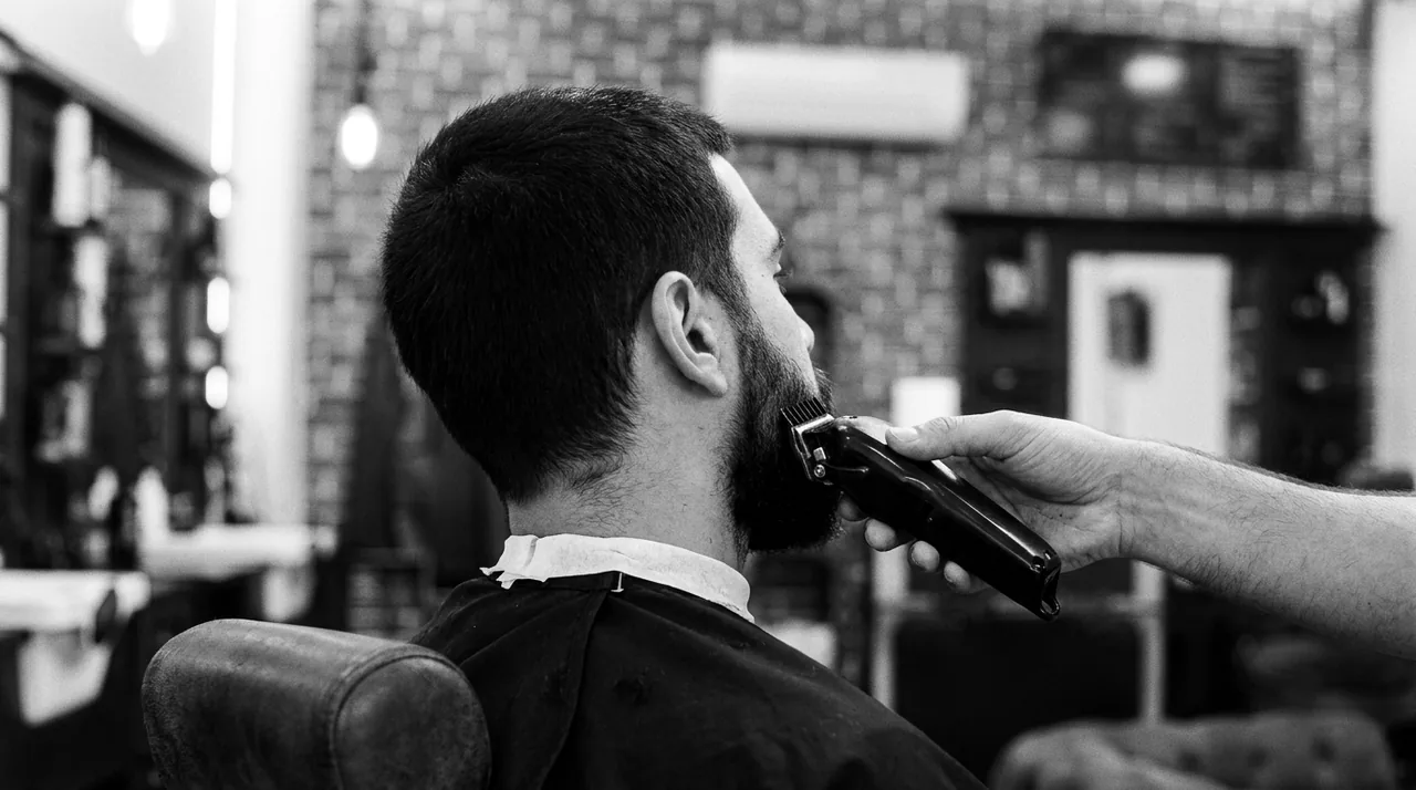 Vue par-dessus l'épaule d'un homme aux cheveux courts : la tondeuse du barbier taille sa barbe
