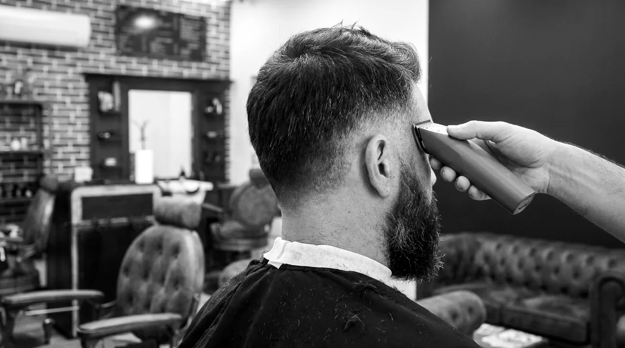 Vue de dos : une tondeuse dégrade le côté du crâne, la barbe encore intacte